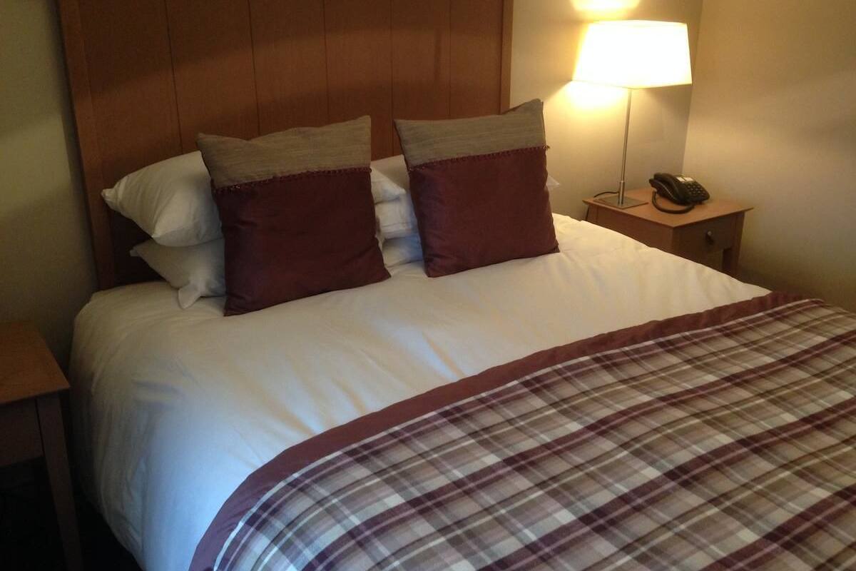 A neatly made bed with white linens and two brown pillows, a plaid blanket, and a bedside lamp on a wooden nightstand.