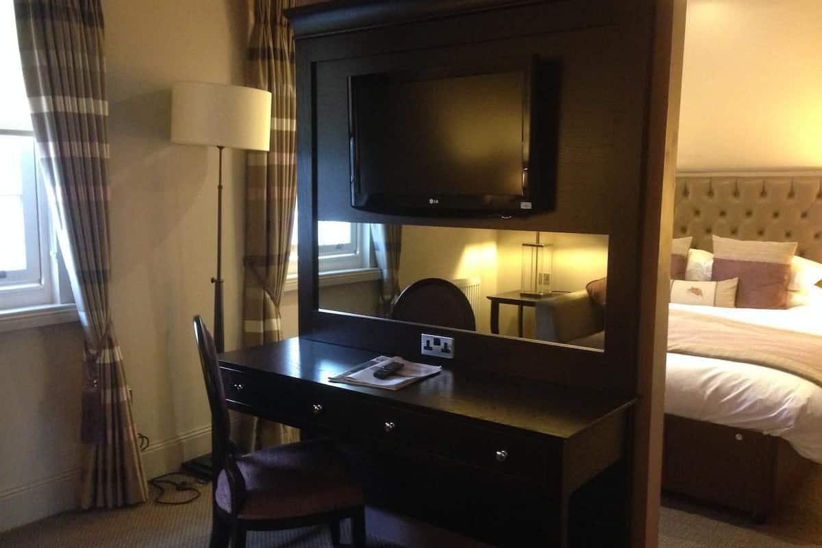 A hotel room with a dark wooden desk, a chair, a flat-screen TV, a lamp, and a bed with pillows.