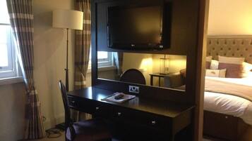 A hotel room with a dark wooden desk, a chair, a flat-screen TV, a lamp, and a bed with pillows.