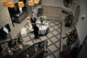 Uma recepção de casamento com um casal sentado à mesa redonda, rodeado por arranjos florais e um chão xadrez.