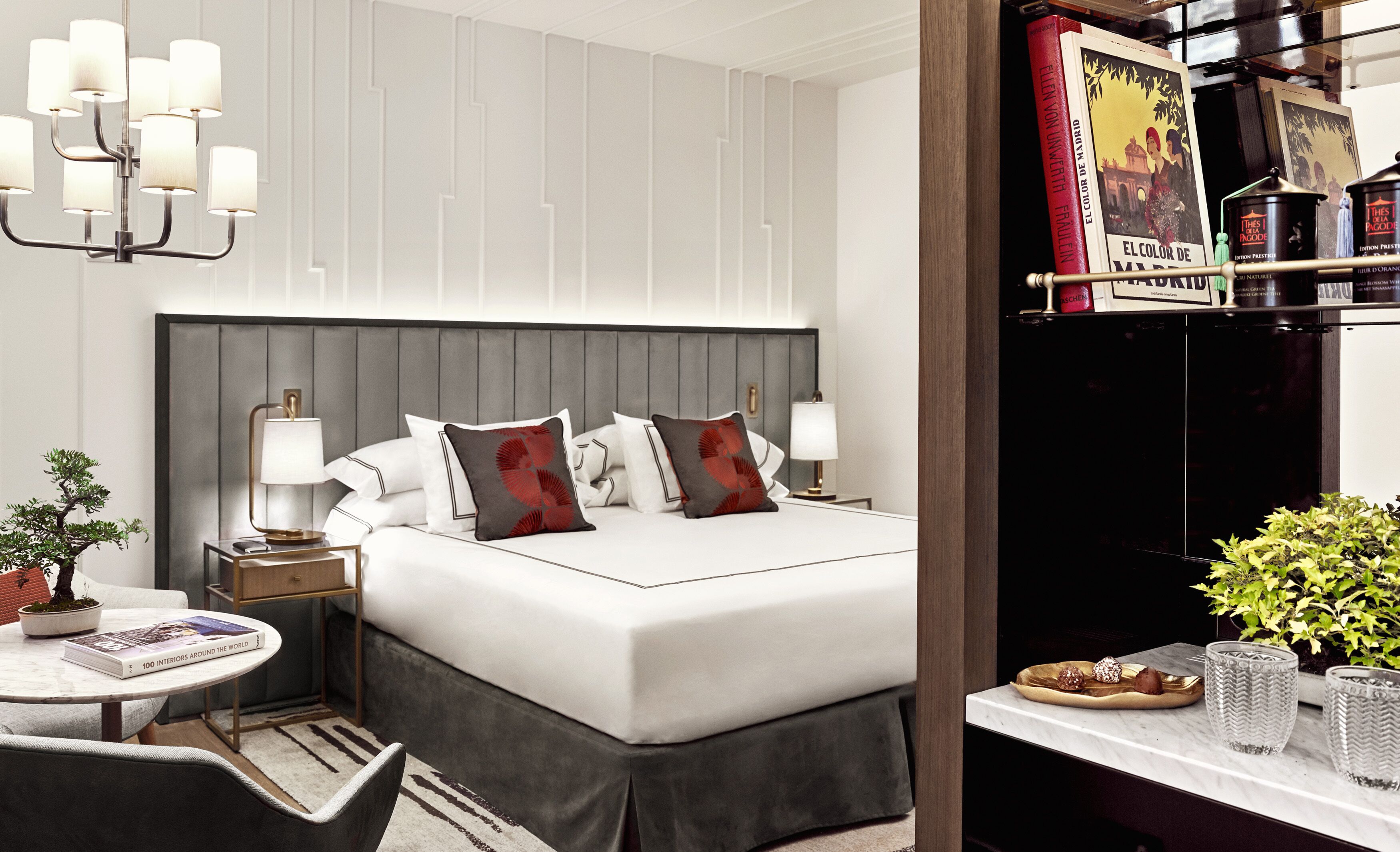 Habitación de hotel con una cama grande, lámparas de noche, una mesita con un libro, una planta en maceta y una estantería llena de libros.