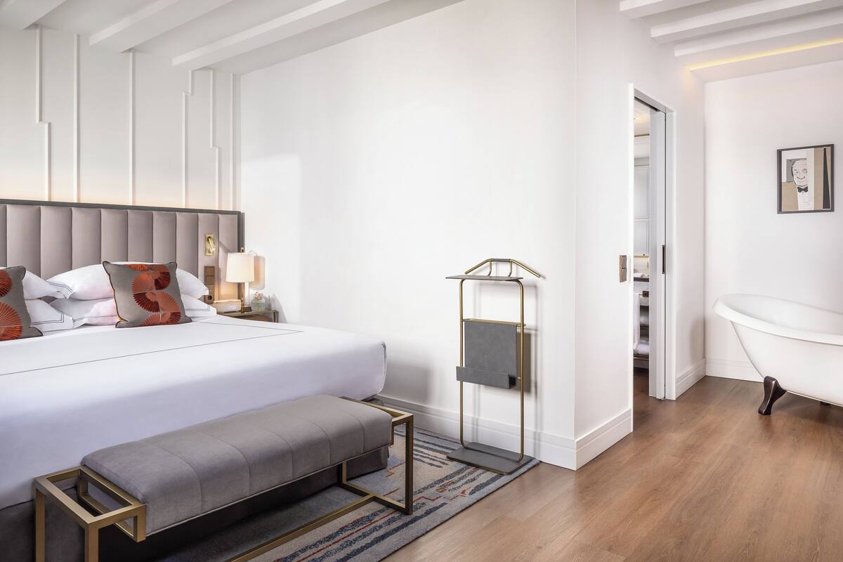 Una habitación de hotel moderna con una cama grande, un banco, una mesita de noche, un televisor montado en la pared y un baño con bañera.
