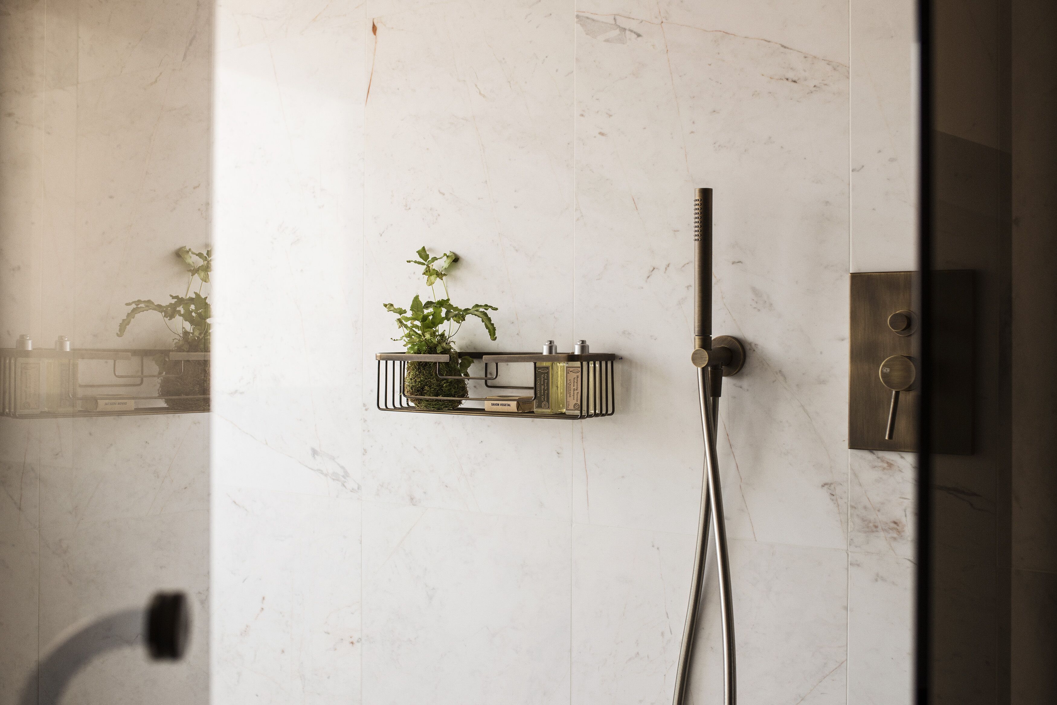 Un baño con pared de mármol, ducha con cabezal de ducha manual y una planta pequeña en una maceta de metal.