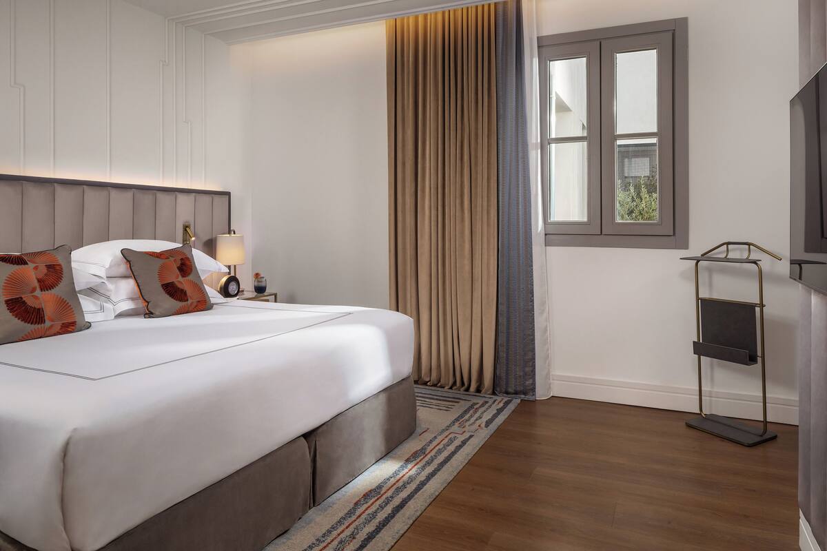Habitación de hotel con una cama grande, una mesita de noche, una lámpara, una ventana con cortinas y un mueble para televisión.