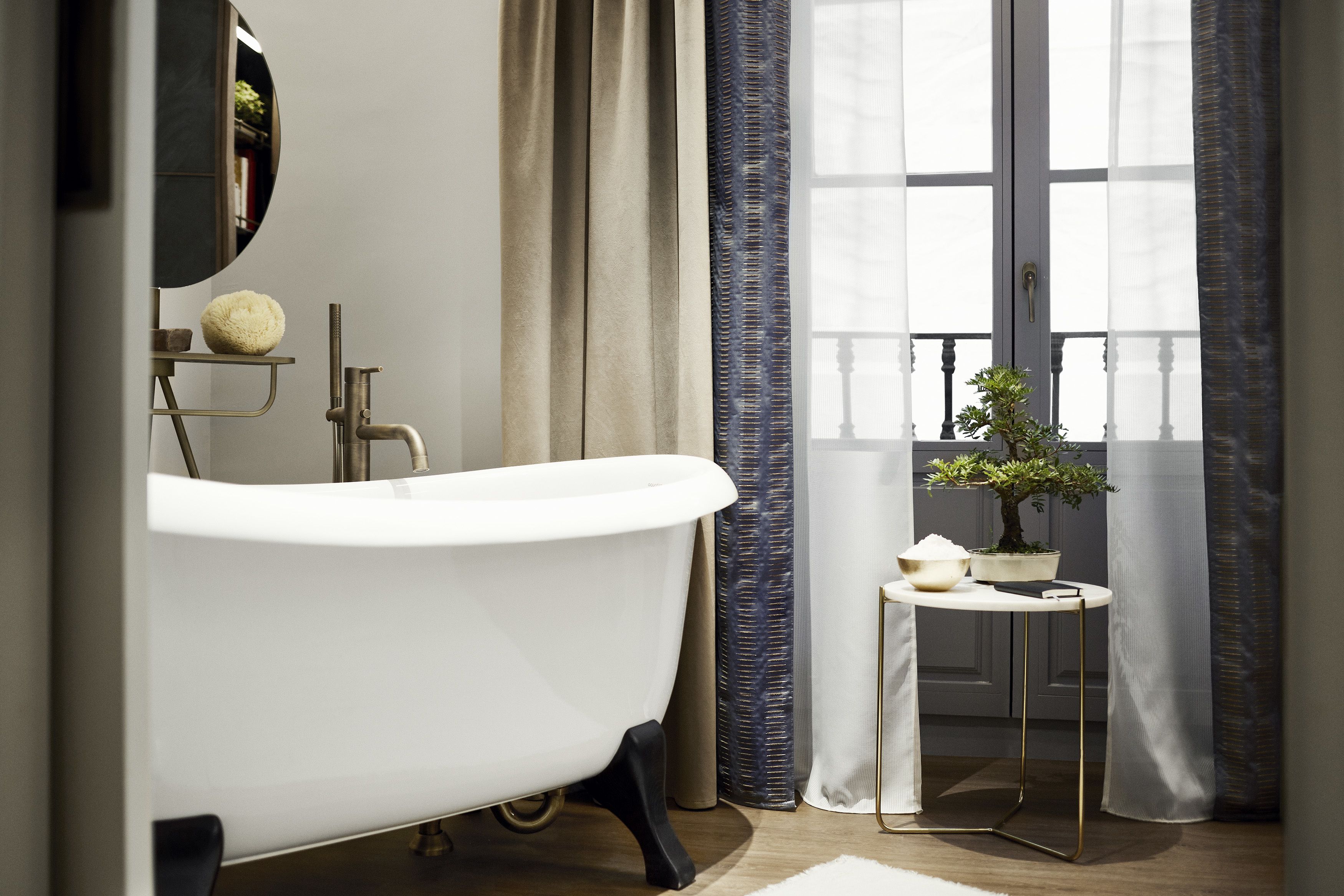 Une salle de bain avec une baignoire sur pieds, une petite table ronde agrémentée d’une plante en pot et une fenêtre avec des rideaux.