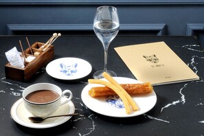 A table with a cup of hot chocolate, a glass of water, a plate with churros, a menu, and a wooden box with chopsticks and sugar cubes. - Gran Hotel Inglés - The Leading Hotels of the World (Madrid)