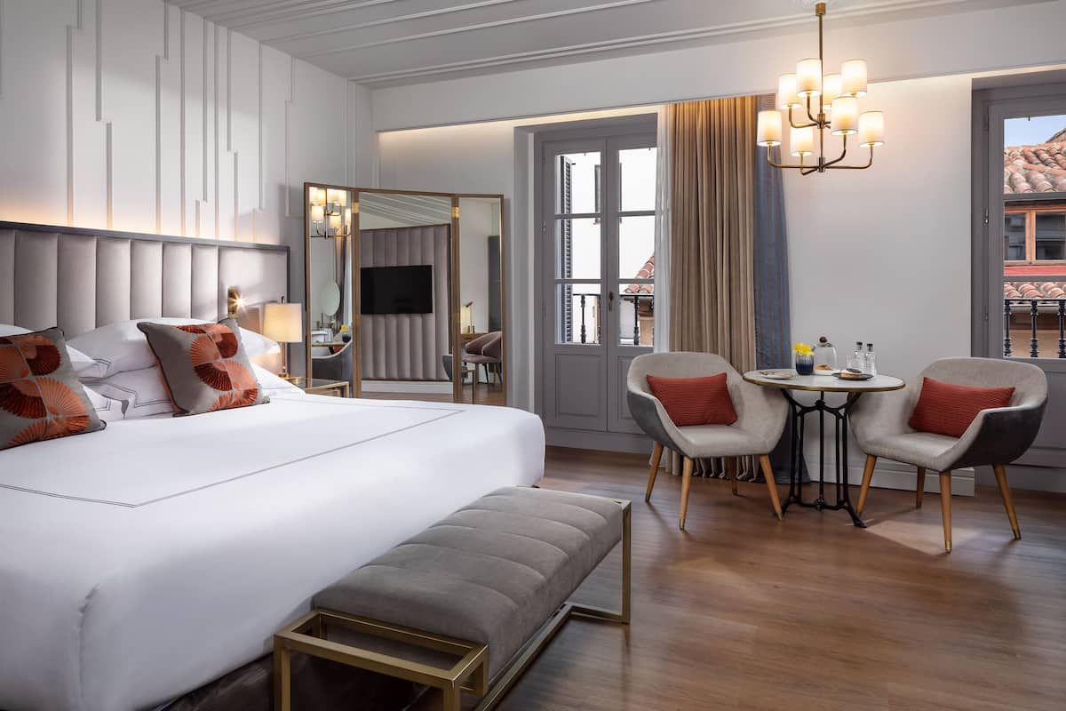 Habitación de hotel con una cama grande, una mesita de noche, una silla, una mesa redonda y una araña.