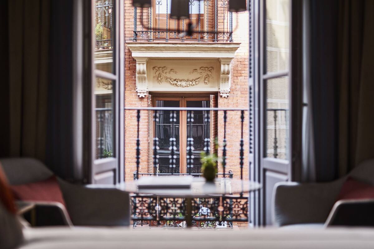 Vista desde un balcón con una mesa y sillas, baranda de hierro decorativa, y fachada de edificio con un balcón y ventanas.