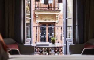 Une vue depuis un balcon, avec une table et des chaises, une balustrade en fer forgé décorative, et la façade d’un immeuble doté d’un balcon et de fenêtres.