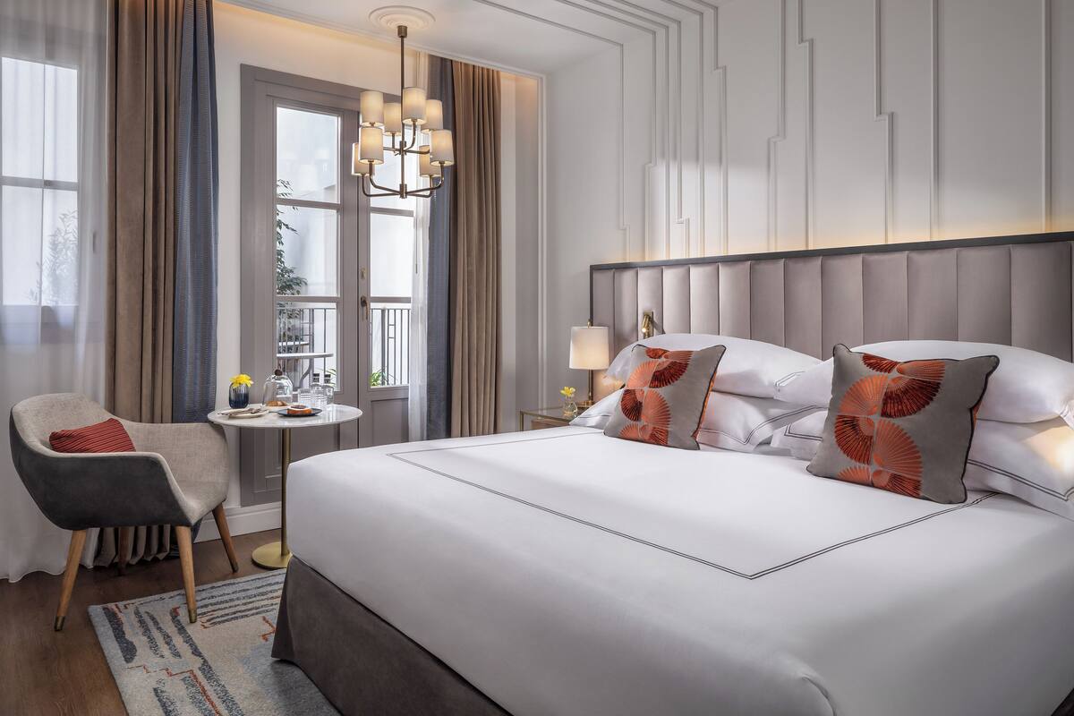Habitación de hotel con una cama grande, una silla, una mesita de noche con lámpara y vistas al exterior por una ventana.