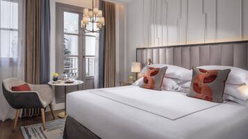Habitación de hotel con una cama grande, una silla, una mesita de noche con lámpara y vistas al exterior por una ventana.