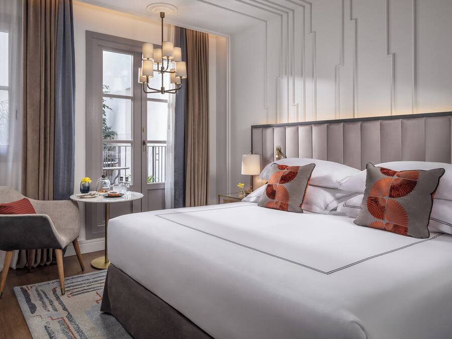 Habitación de hotel con una cama grande, una silla, una mesita de noche con lámpara y vistas al exterior por una ventana.