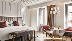 A modern hotel room with a large bed, bedside tables, a seating area with a table and chairs, and a chandelier.