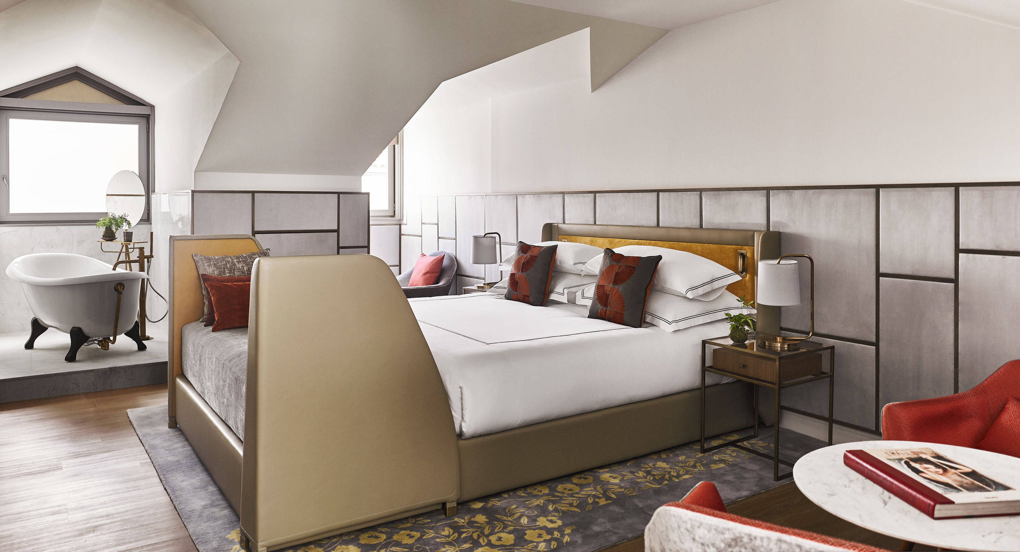 Una habitación de hotel moderna con una cama grande, un área de descanso con una silla roja, una mesa redonda, una mesita de noche con una lámpara y una bañera.