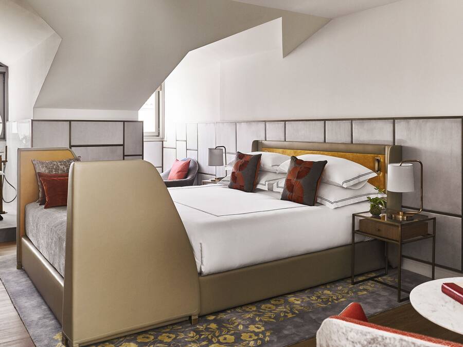 Una habitación de hotel moderna con una cama grande, un área de descanso con una silla roja, una mesa redonda, una mesita de noche con una lámpara y una bañera.