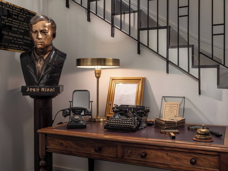 Un escritorio con un busto de José Rizal, una máquina de escribir antigua, una lámpara y un menú enmarcado.