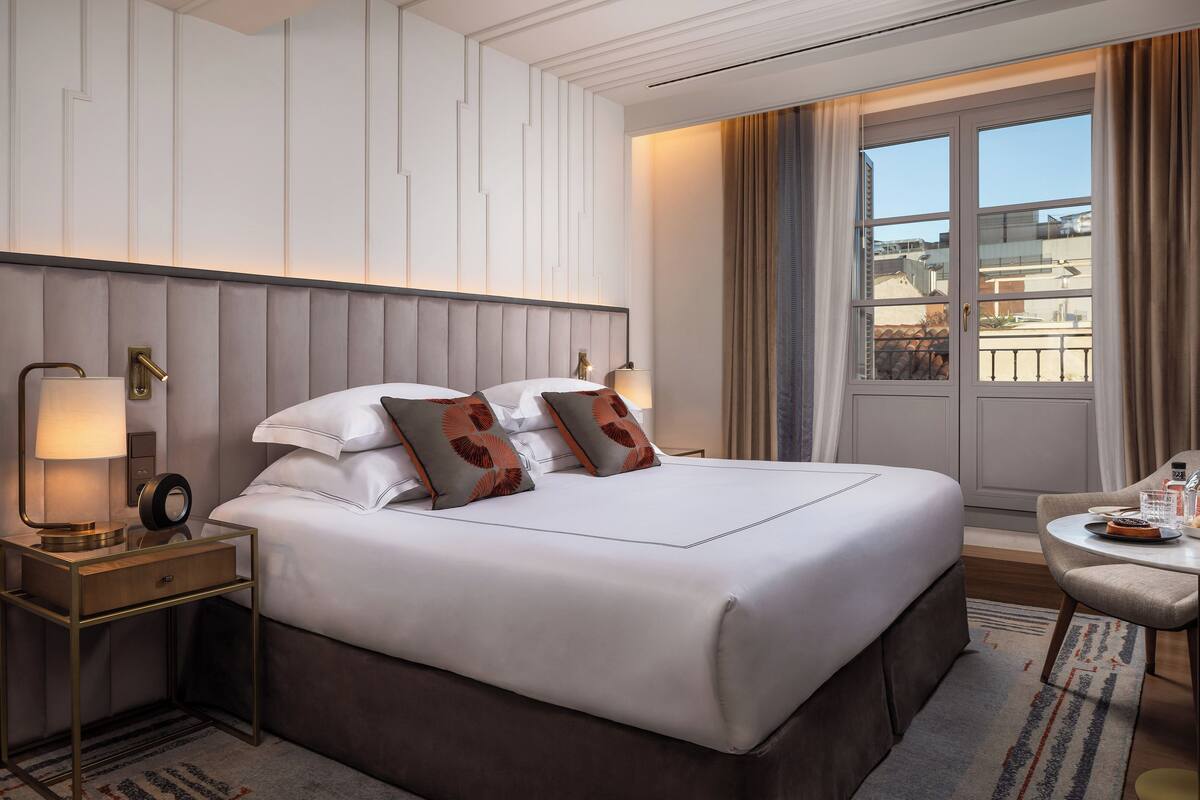 Habitación de hotel con una cama grande, una mesita de noche con lámpara, una pequeña mesa con silla y vistas al exterior a través de una ventana.