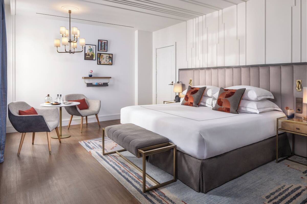 Una habitación de hotel moderna con una cama grande, mesitas de noche, una pequeña mesa redonda, un banco y una araña.