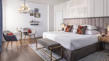 A modern hotel room with a large bed, bedside tables, a small round table, a bench, and a chandelier.
