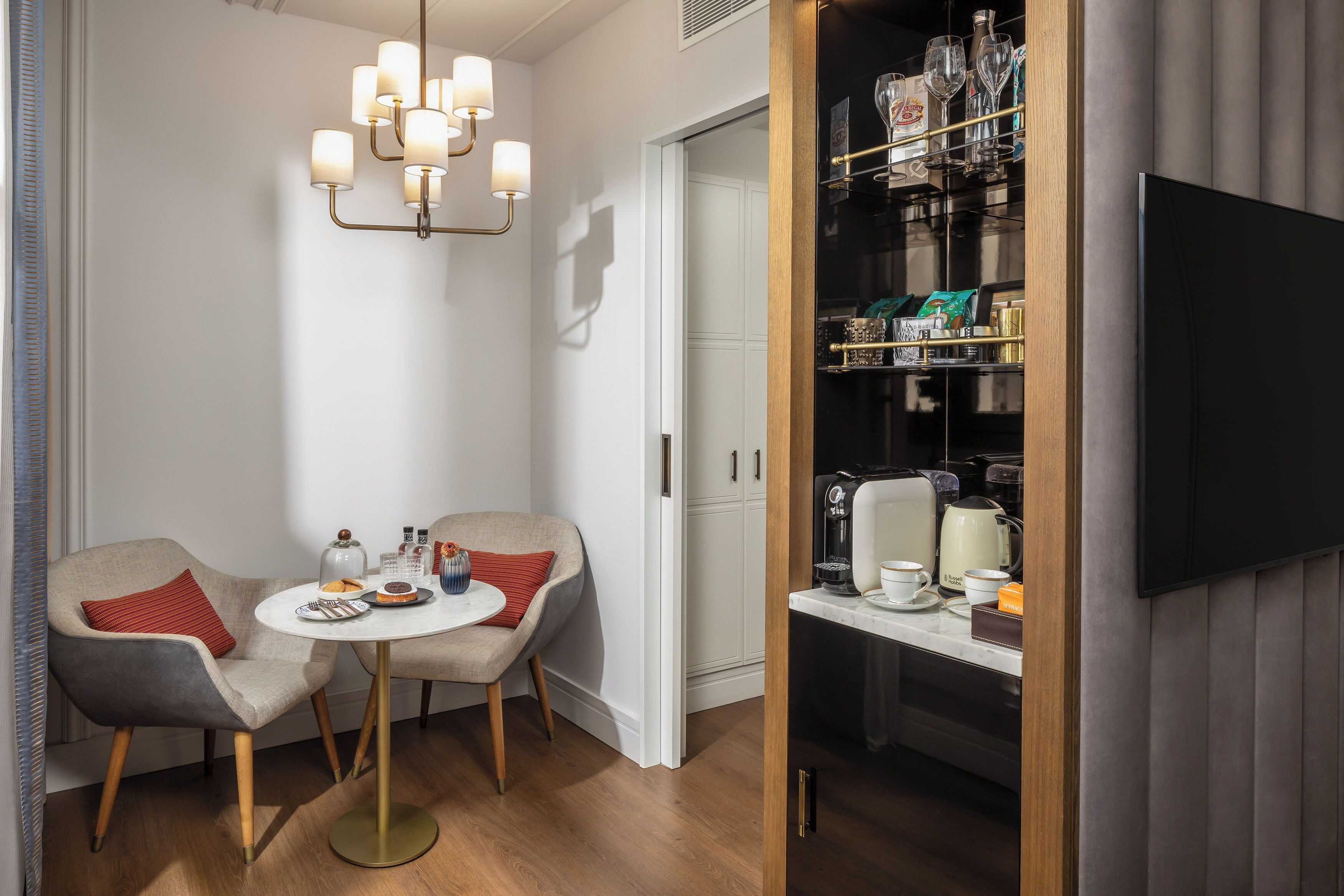 A modern hotel room with a dining area featuring two chairs, a round table, and a chandelier. A minibar is visible with various items and a coffee machine.