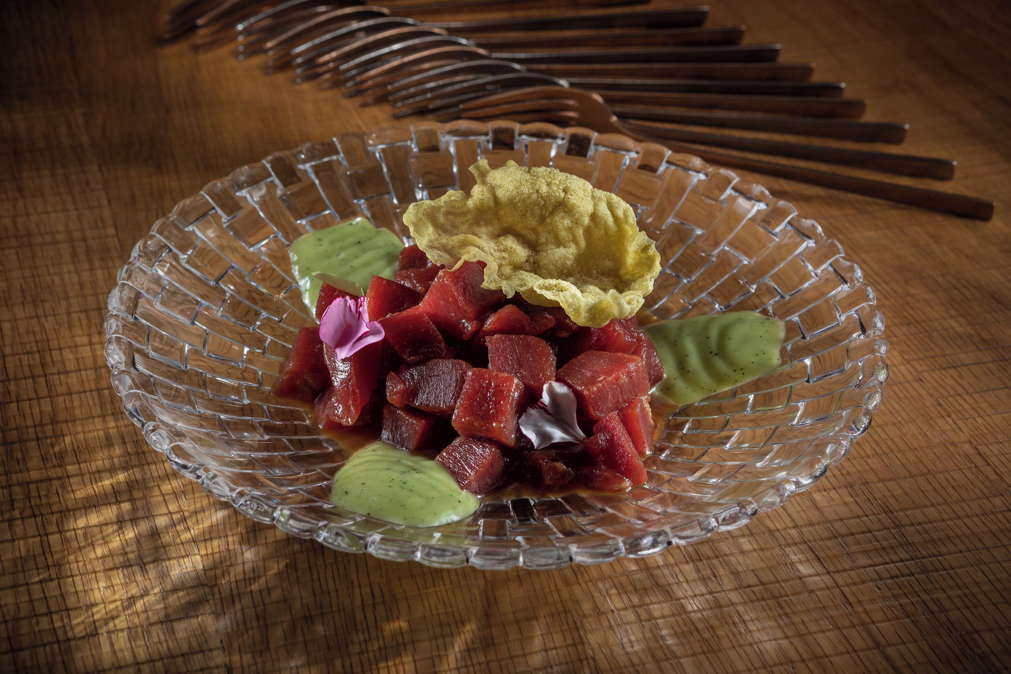 A glass bowl with diced red fruit, green leaves, and a yellow item on a wooden surface.