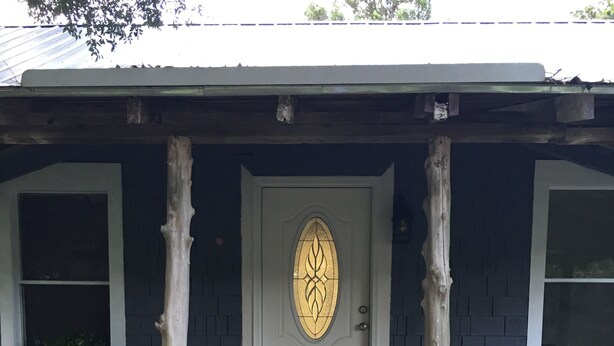 A wooden porch with a white door featuring a decorative glass panel, flanked by two wooden railings and potted plants.