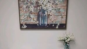 A white cabinet with three drawers and two doors, a vase with white flowers, and a painting of a vase with flowers on the wall above.