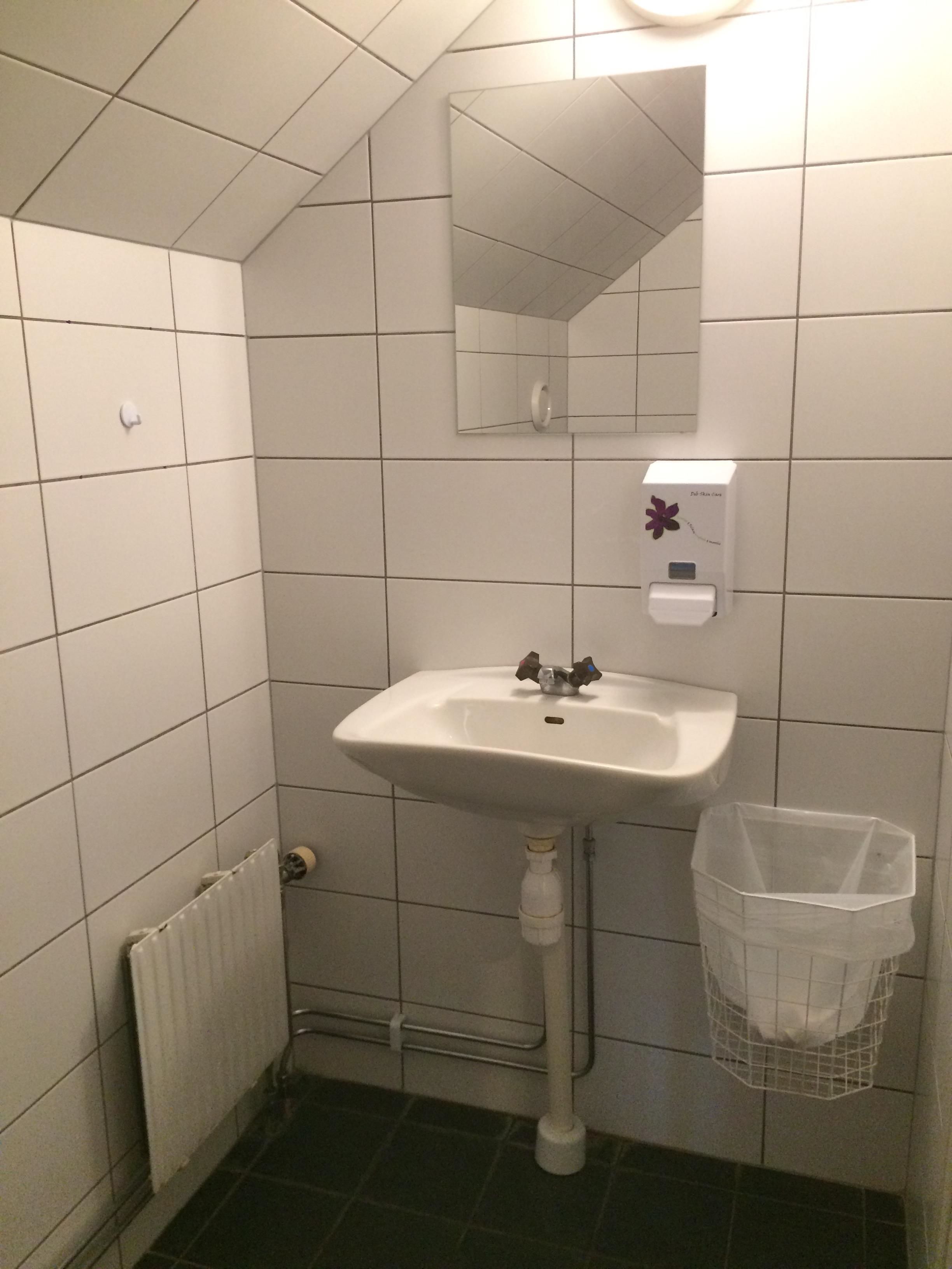 Un baño pequeño con azulejos blancos, un lavamanos, un espejo, un secador de manos y un cubo de basura.