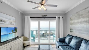 Un salon équipé d’une télévision, d’un ventilateur de plafond, d’un canapé et offrant une vue sur la plage grâce à de grandes portes coulissantes.