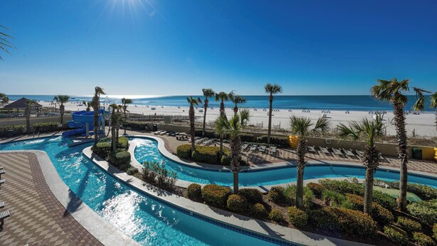 A pool area with multiple water features, palm trees, and a beach in the background. - Phoenix West 3BR! LOW FLOOR! (Orange Beach)