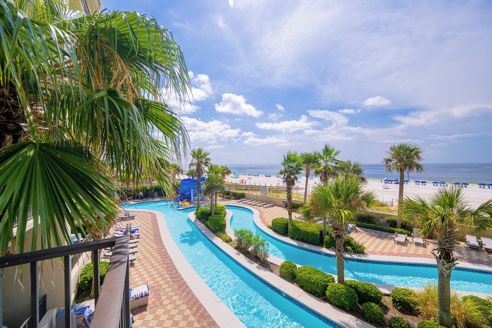 A resort pool area with a view of the beach, lounge chairs, and palm trees.