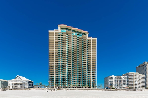Een hoog hotel met meerdere balkons, gelegen aan een strand met andere gebouwen in de buurt.