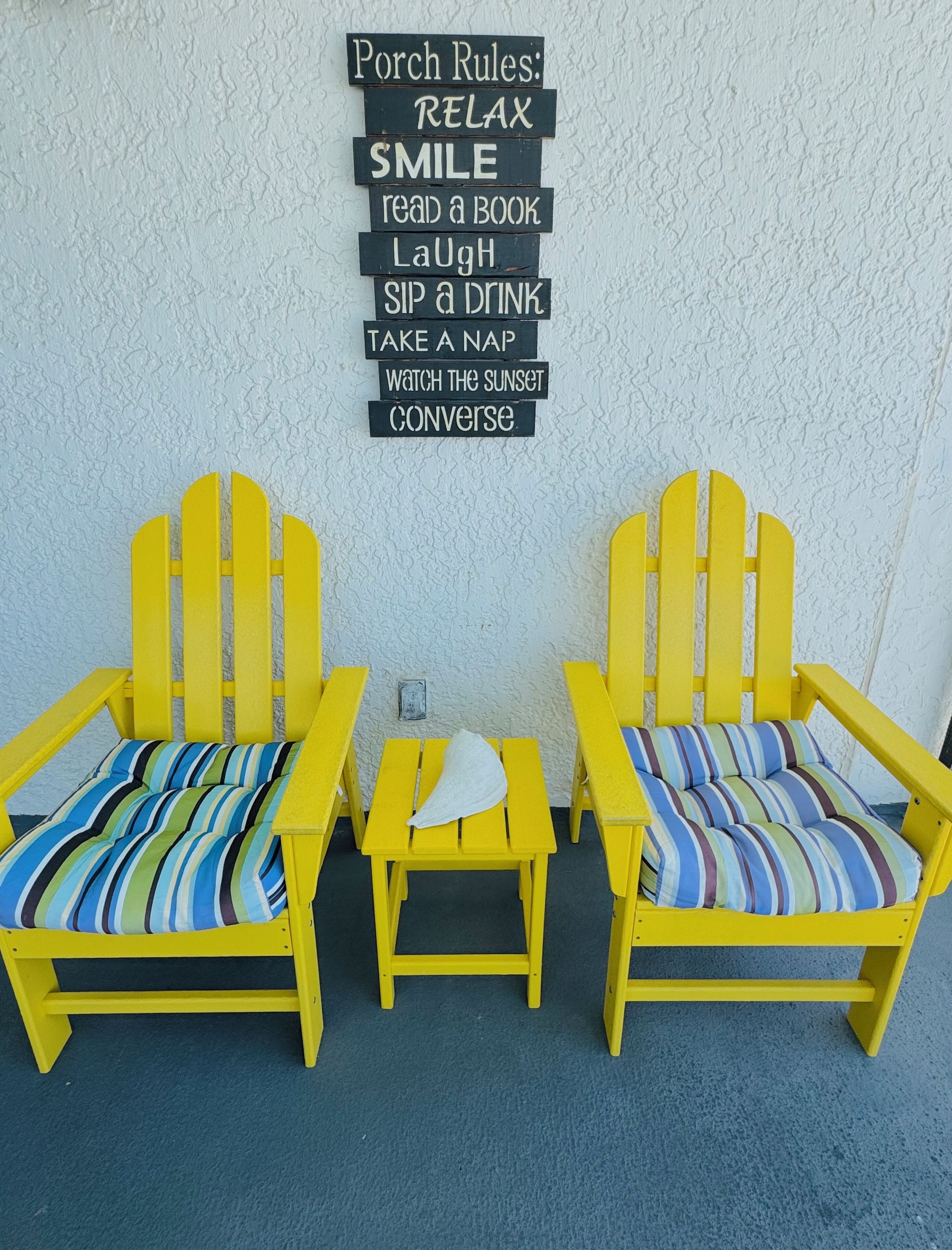 Zwei gelbe Adirondack-Stühle mit gestreiften Kissen, ein kleiner Tisch und ein an der Wand befestigtes Schild mit der Aufschrift 'Verhaltensregeln auf der Veranda', das Aktivitäten wie Lesen und Sonnenuntergang beobachten auflistet.