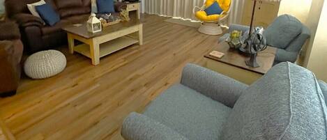 Un salon avec un canapé marron, un canapé bleu, une table basse en bois et une chaise jaune. Les murs sont décorés et il y a un ventilateur de plafond.