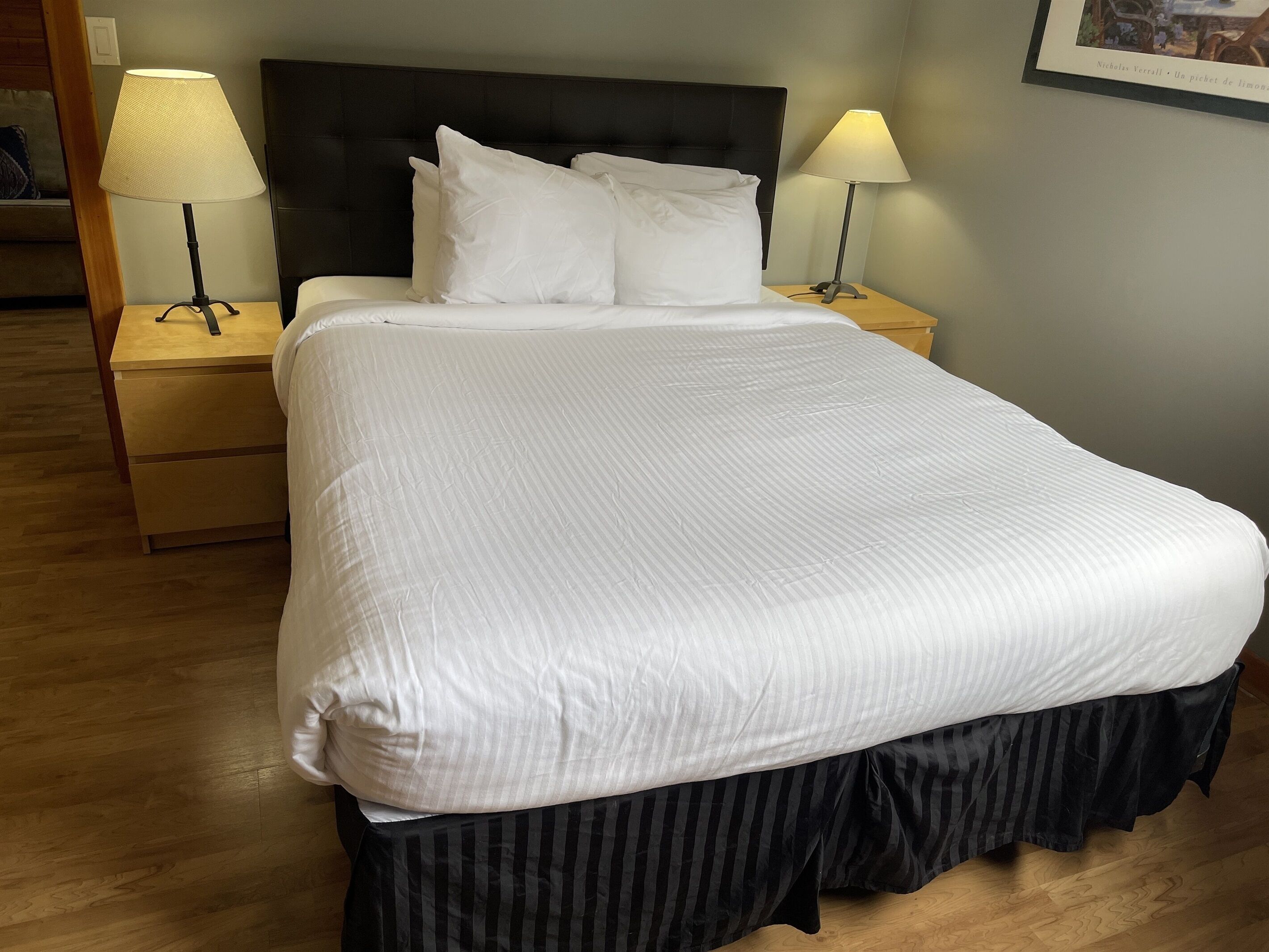 Una cama bien hecha con sábanas blancas y falda a rayas, dos lámparas de noche y una mesita de madera.