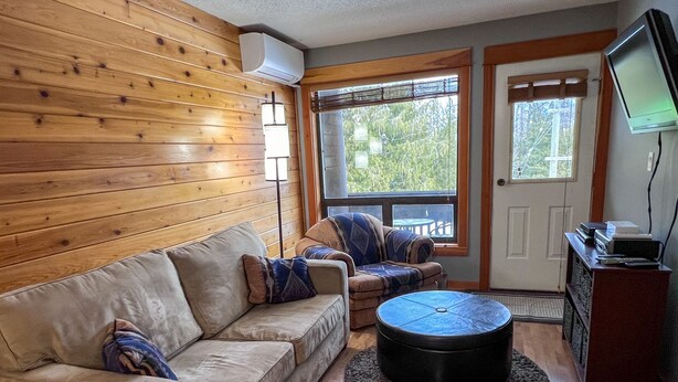 A cozy living room with a wooden wall, a beige sofa, a patterned armchair, a round coffee table, and a television on a wooden stand.