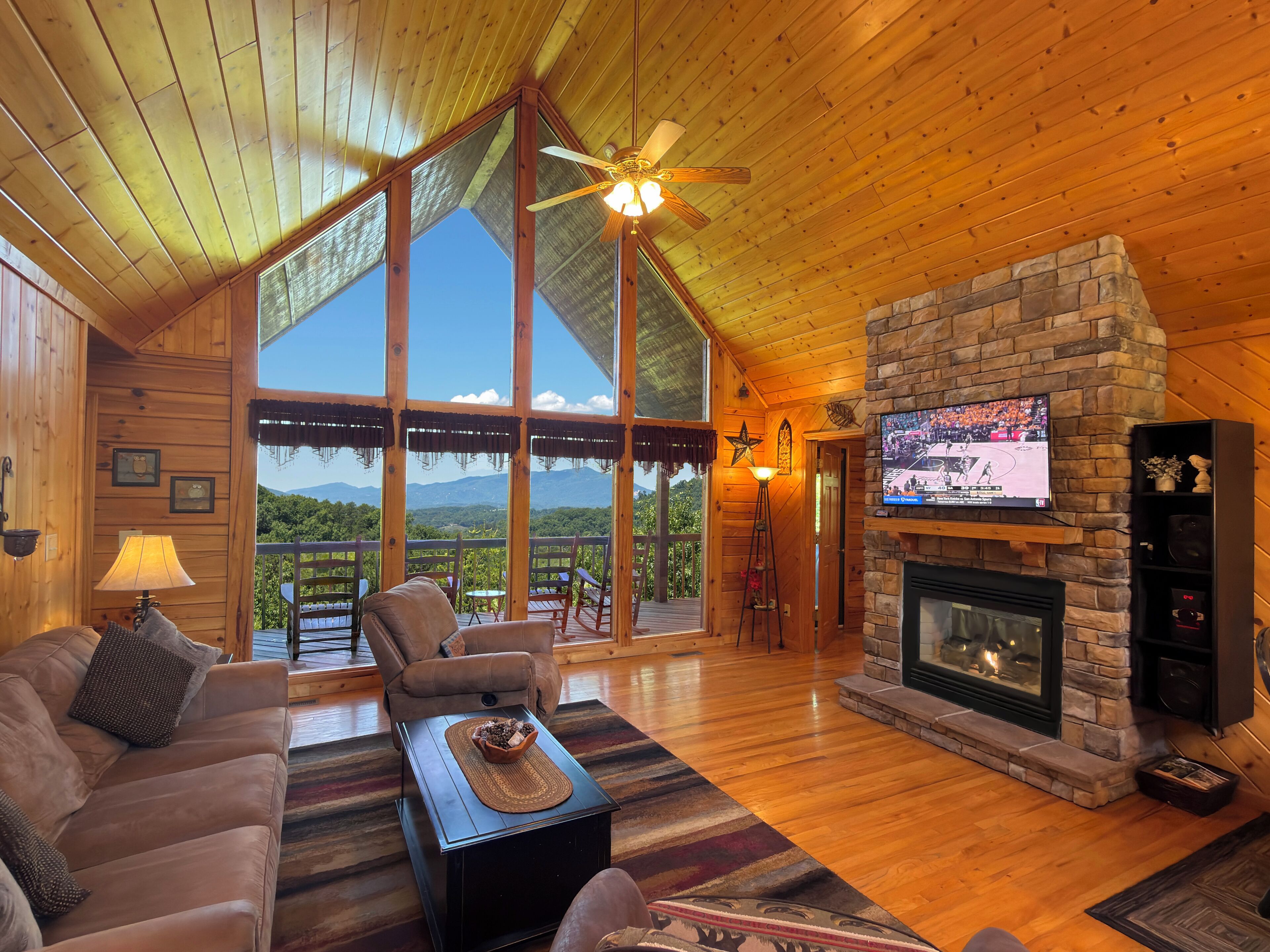 A cozy cabin interior with a wooden ceiling, a stone fireplace, a television, and a large window overlooking a mountainous landscape.