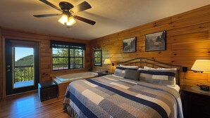 Ein Schlafzimmer in einer Holzhütte mit einem großen Bett, einem Deckenventilator, einer Badewanne und einem Balkon mit Blick auf einen bewaldeten Hügel.