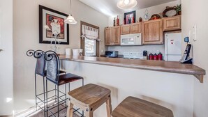 Cocina compacta con armarios de madera, microondas, refrigerador y una barra con taburetes.