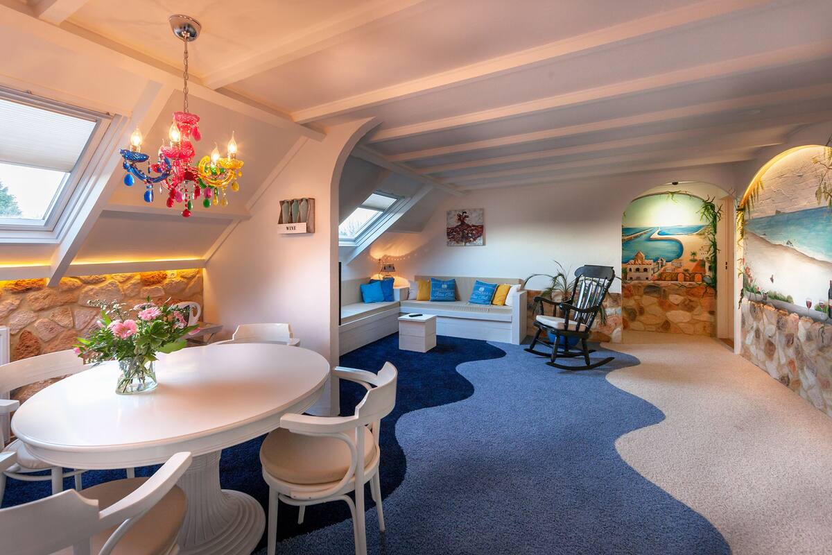 A cozy room with a round dining table, white chairs, a blue carpet, a stone wall, a wooden bench, and a colorful chandelier.
