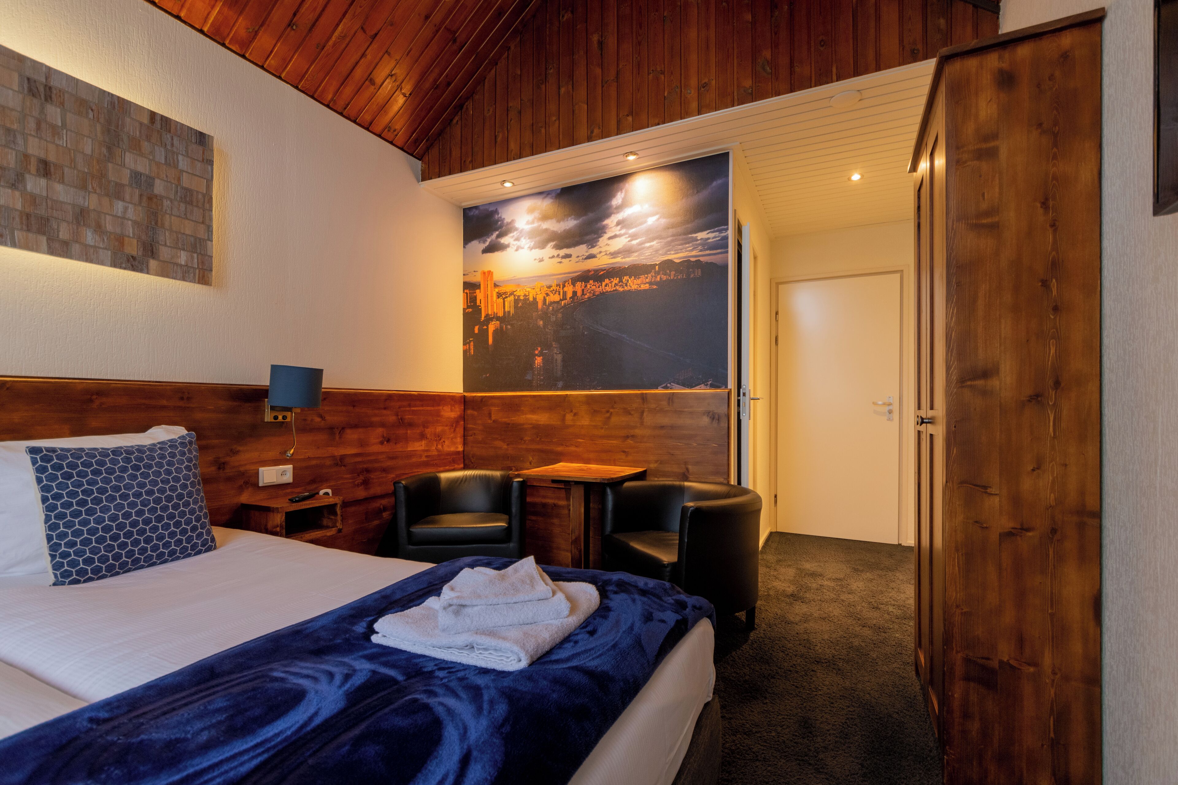 Una camera d'albergo con un letto, un'area salotto con un tavolo, un grande poster appeso al muro e un armadio.
