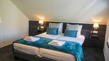 Cama doble con colcha verde aguamarina, dos almohadas estampadas y dos toallas blancas dobladas sobre la cama. Hay dos mesitas de noche con lámparas, un cabecero oscuro y una pared de color claro.