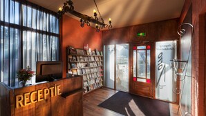 Un espace d’accueil avec un bureau en bois portant l’inscription « RÉCEPTION », une bibliothèque contenant divers objets et une grande fenêtre laissant entrer la lumière naturelle.