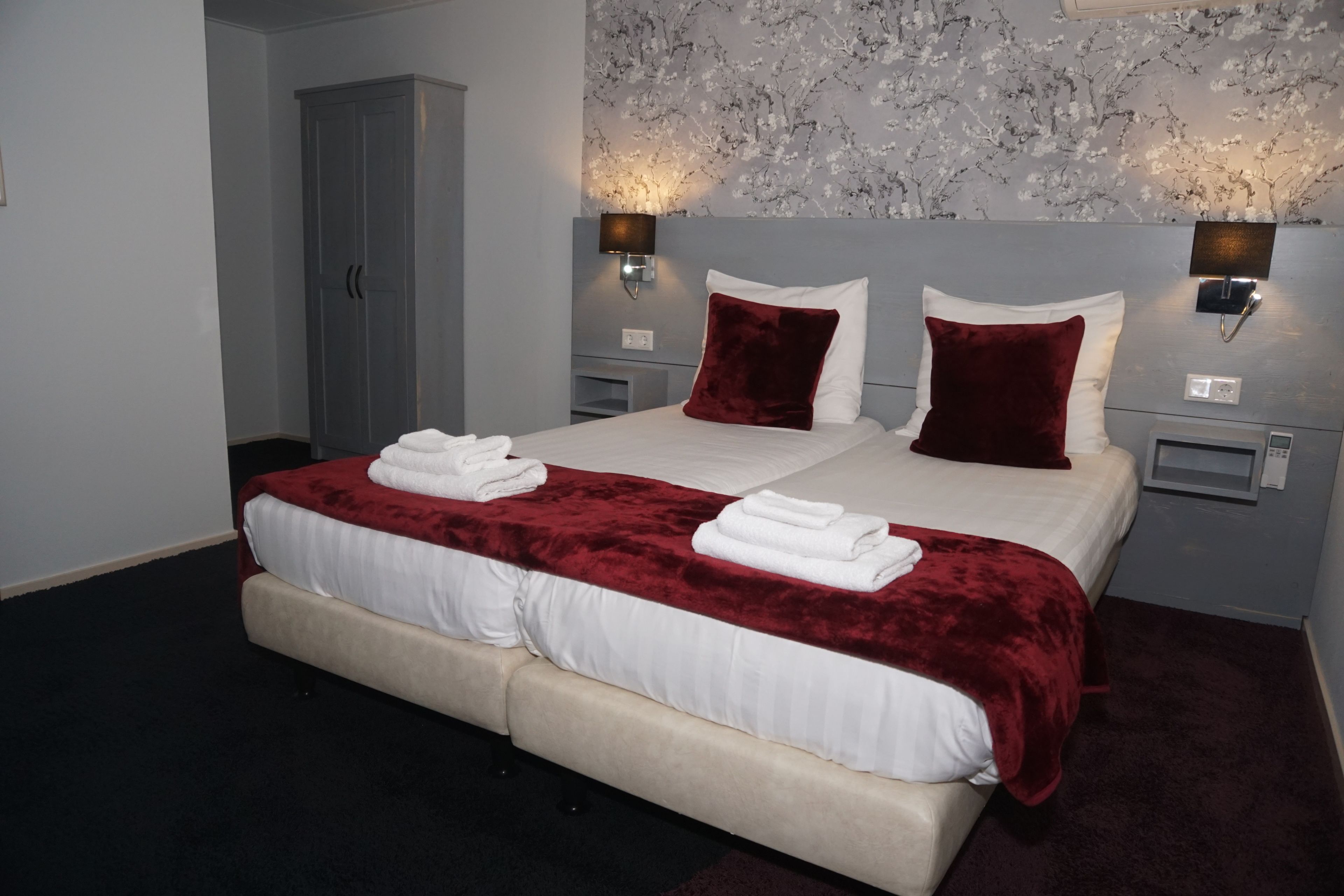 A double bed with white linens and a red velvet throw, two wall-mounted lamps, and a floral-patterned wall.