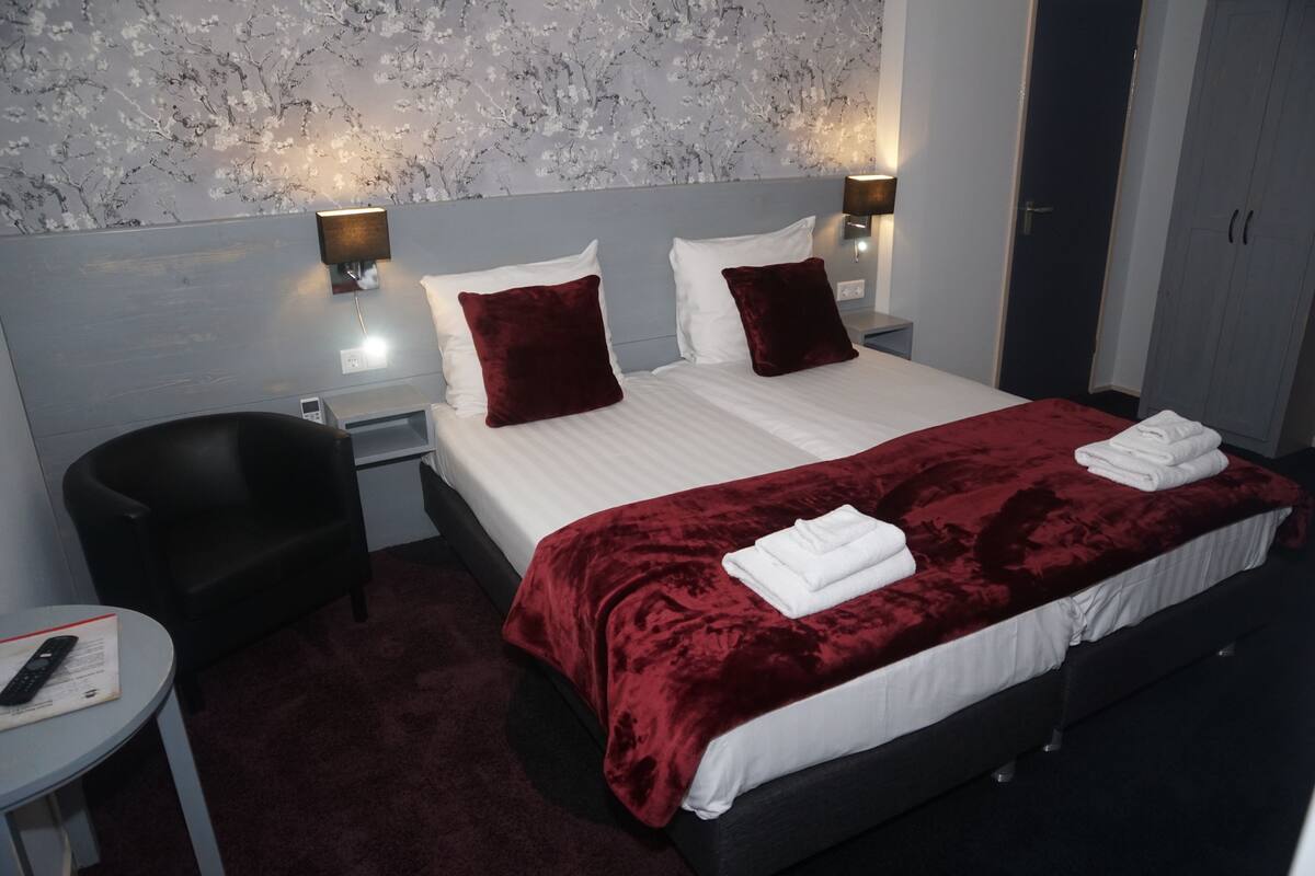 A hotel room with a large bed, two pillows, a red blanket, a black chair, a small table with a remote control, and a nightstand with a lamp.
