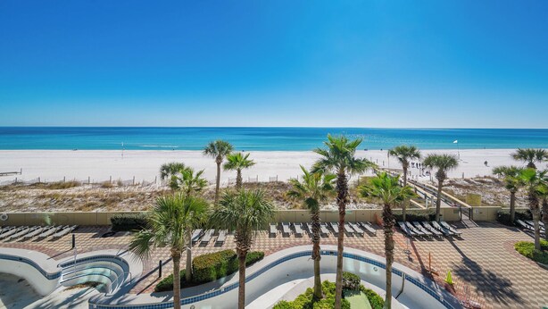 Vista a la playa con palmeras, una zona de piscina y un cielo azul despejado.