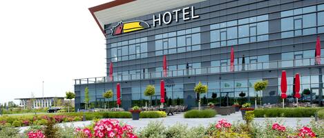 A modern hotel with a large glass facade, outdoor seating area with red umbrellas, and a well-maintained garden with pink flowers.