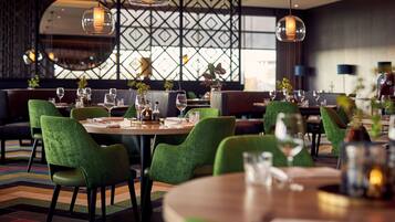 Un restaurante con mesas redondas, sillas tapizadas en verde y lámparas colgantes.