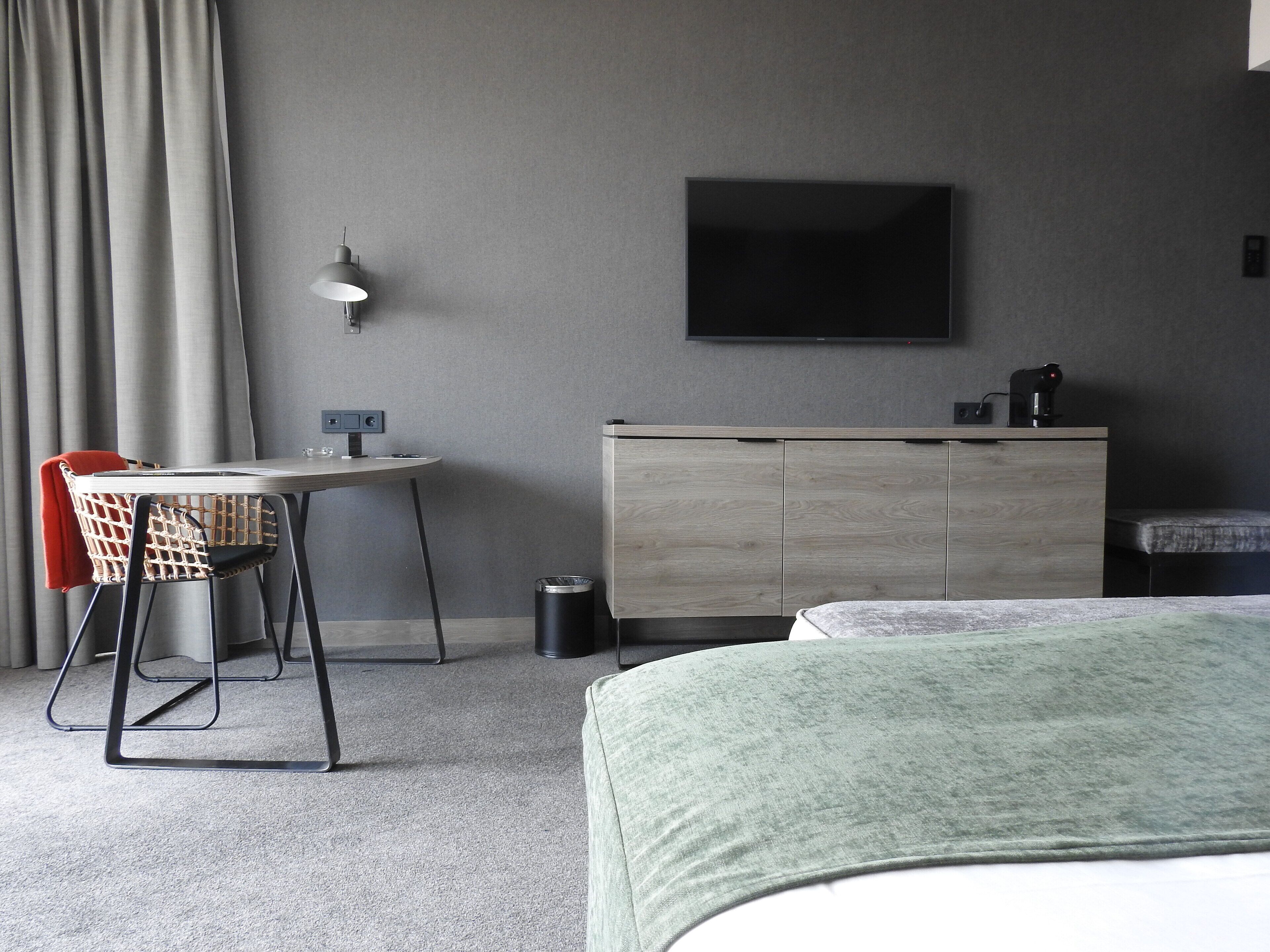 A hotel room with a bed, a desk with a chair, a television mounted on the wall, and a small cabinet.
