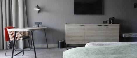 Una habitación de hotel con una cama, un escritorio con silla, un televisor montado en la pared y un pequeño armario.